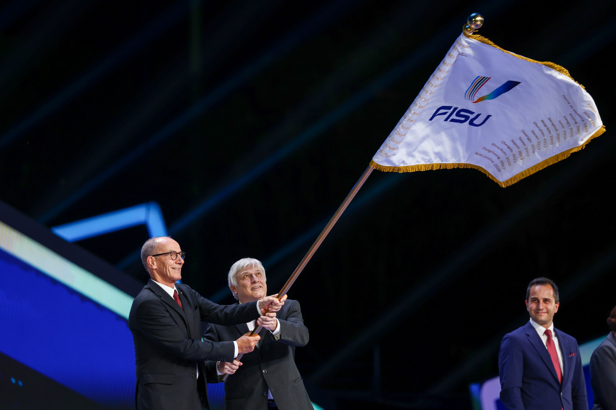 Mahmut Özdemir, right, member of the German Bundestag, watches on as he prepares to receive the FISU flag from FISU Acting President Leonz Eder and chief executive and secretary general Eric Saintrond ©Chengdu 2021