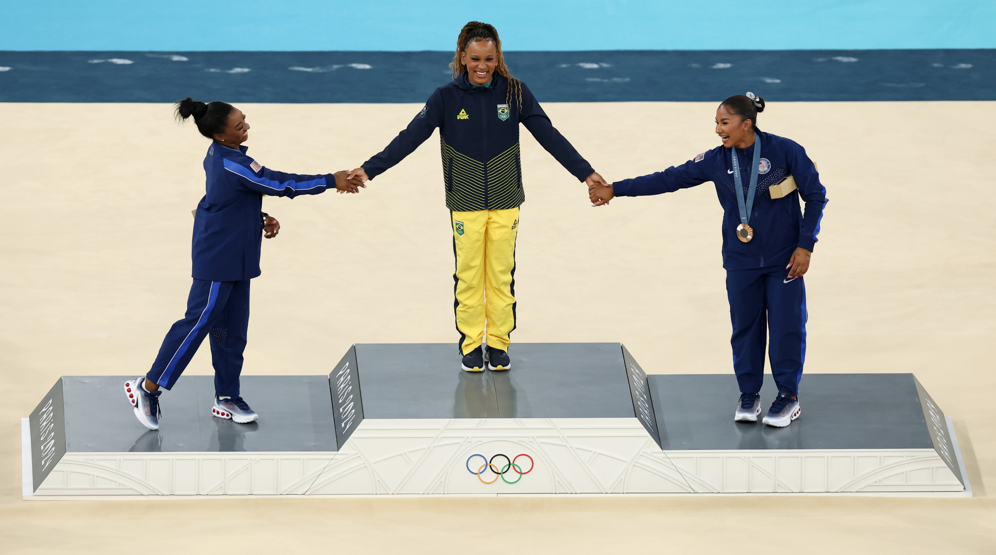 Gold medalist Rebeca Andrade of Brazil holds hands with Simone Biles and Jordan Chiles of Team United States on the podium at the Artistic Gymnastics Women's Floor Exercise Medal Ceremony at the Paris 2024 Olympic Games. GETTY IMAGES