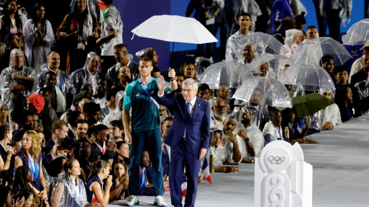 Volunteer shields Thomas Bach from the rain, allowing the IOC President to greet. GETTY IMAGES